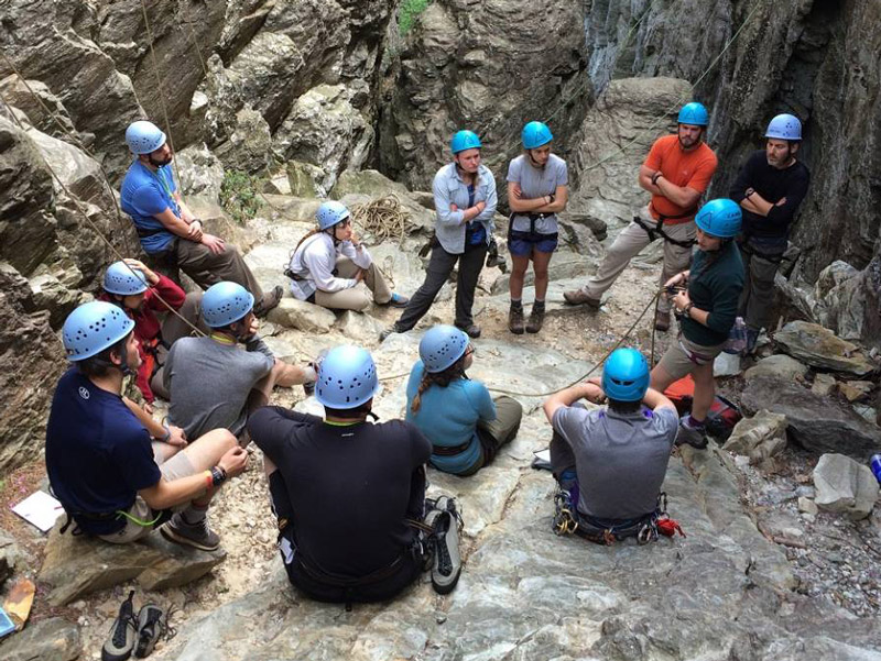 students wearing helmets sit in a circle and listen to rock climbing instructions